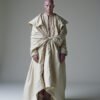 A figure in a flowing beige garment with oversized sleeves and a waist knot, standing confidently against a minimalist background, evoking themes of strength, identity, and self-expression.