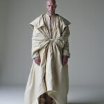 A figure in a flowing beige garment with oversized sleeves and a waist knot, standing confidently against a minimalist background, evoking themes of strength, identity, and self-expression.